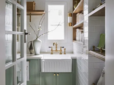 A butler's pantry with white subway tiled walls, a sizable fluted fireclay sink, hardware in Satin Brass and joinery finished in a mint green colour. Timber open shelving allows owners to display trays and dinnerware, plus containers filled with ingredients. The tiled flooring has a star pattern.
