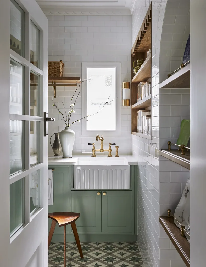 A butler's pantry with white subway tiled walls, a sizable fluted fireclay sink, hardware in Satin Brass and joinery finished in a mint colour. Timber open shelving allows owners to display trays and dinnerware, plus containers filled with ingredients. The tiled flooring has a star pattern.
