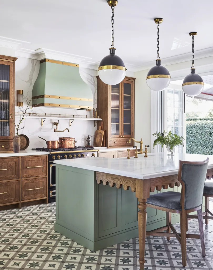 A green kitchen that was renovated with English style features to suit the 1921 build. A mint green finish is used on the kitchen island and rangehood, with three pendant lights suspended above the island and wooden joinery against the wall. Calacatta oro stone is the benchtop and splashback, which reaches all the way to the white ceiling. A La Cornue oven is opposite the island. The tiled flooring has a star pattern.