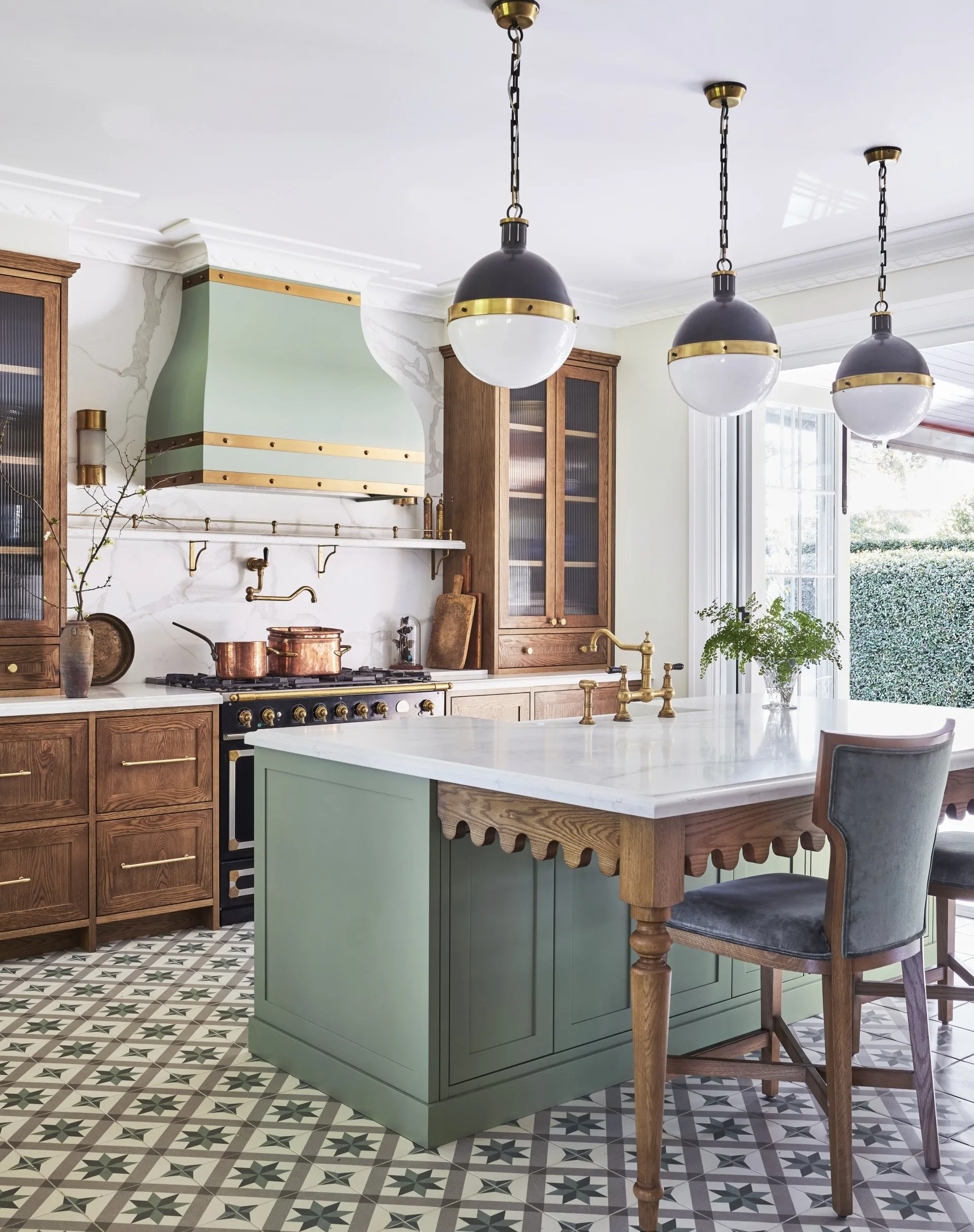 A green kitchen that was renovated with English style features to suit the 1921 build. A mint green finish is used on the kitchen island and rangehood, with three pendant lights suspended above the island and wooden joinery against the wall. Calacatta oro stone is the benchtop and splashback, which reaches all the way to the white ceiling. A La Cornue oven is opposite the island. The tiled flooring has a star pattern.