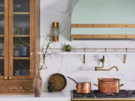 A green kitchen that was renovated with English style features to suit the 1921 build. A mint green finish is used on the kitchen rangehood, above the La Cornue matte black oven and stovetop. Wooden joinery is against the wall on either side. Calacatta oro stone is the benchtop and splashback, which reaches all the way to the white ceiling. Copper pots are placed on the stovetop and a brass pot filler is positioned above these.