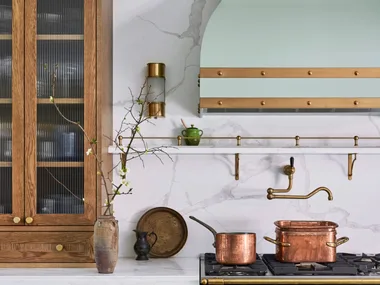 A green kitchen that was renovated with English style features to suit the 1921 build. A mint green finish is used on the kitchen rangehood, above the La Cornue matte black oven and stovetop. Wooden joinery is against the wall on either side. Calacatta oro stone is the benchtop and splashback, which reaches all the way to the white ceiling. Copper pots are placed on the stovetop and a brass pot filler is positioned above these.