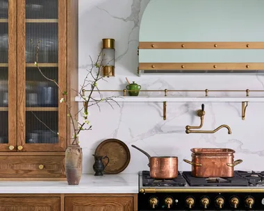A green kitchen that was renovated with English style features to suit the 1921 build. A mint green finish is used on the kitchen rangehood, above the La Cornue matte black oven and stovetop. Wooden joinery is against the wall on either side. Calacatta oro stone is the benchtop and splashback, which reaches all the way to the white ceiling. Copper pots are placed on the stovetop and a brass pot filler is positioned above these.