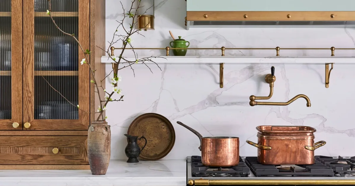 A green kitchen that was renovated with English style features to suit the 1921 build. A mint green finish is used on the kitchen rangehood, above the La Cornue matte black oven and stovetop. Wooden joinery is against the wall on either side. Calacatta oro stone is the benchtop and splashback, which reaches all the way to the white ceiling. Copper pots are placed on the stovetop and a brass pot filler is positioned above these.