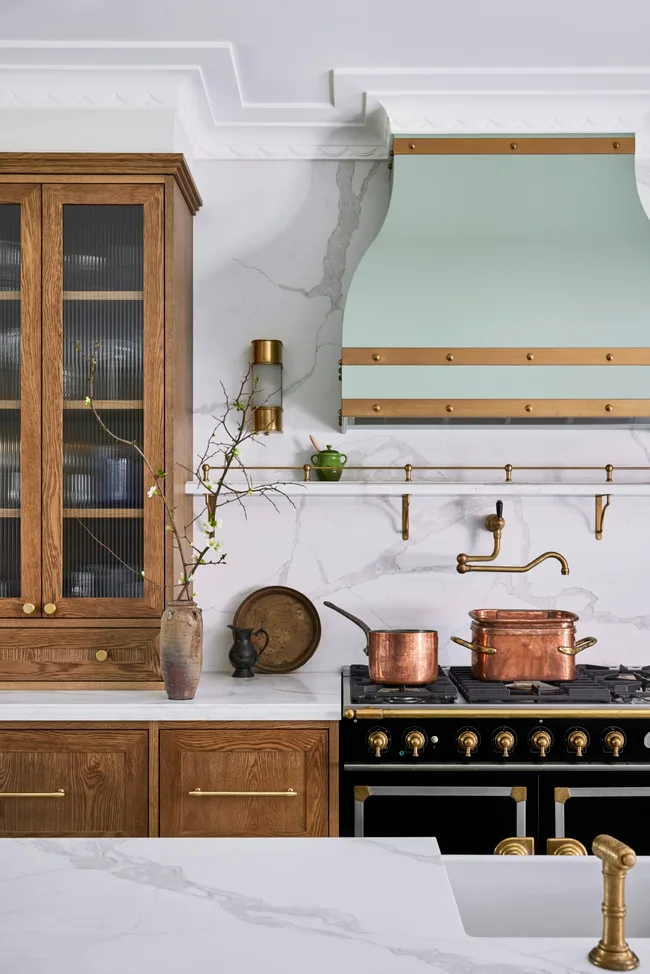 A green kitchen that was renovated with English style features to suit the 1921 build. A mint green finish is used on the kitchen rangehood, above the La Cornue matte black oven and stovetop. Wooden joinery is against the wall on either side. Calacatta oro stone is the benchtop and splashback, which reaches all the way to the white ceiling. Copper pots are placed on the stovetop and a brass pot filler is positioned above these.