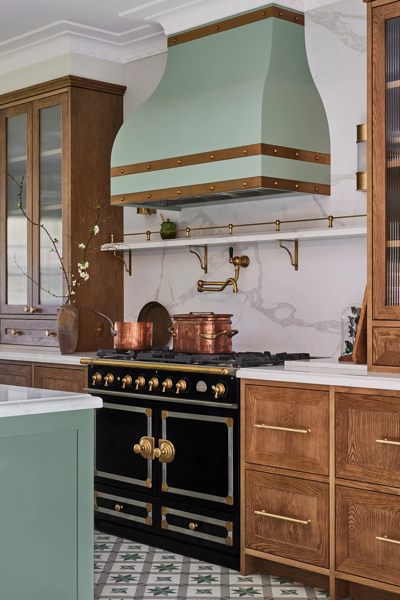 A green kitchen that was renovated with English style features to suit the 1921 build. A mint green finish is used on the kitchen island and rangehood. Wooden joinery is against the wall. Calacatta oro stone is the benchtop and splashback, which reaches all the way to the white ceiling. A La Cornue oven is opposite the island.