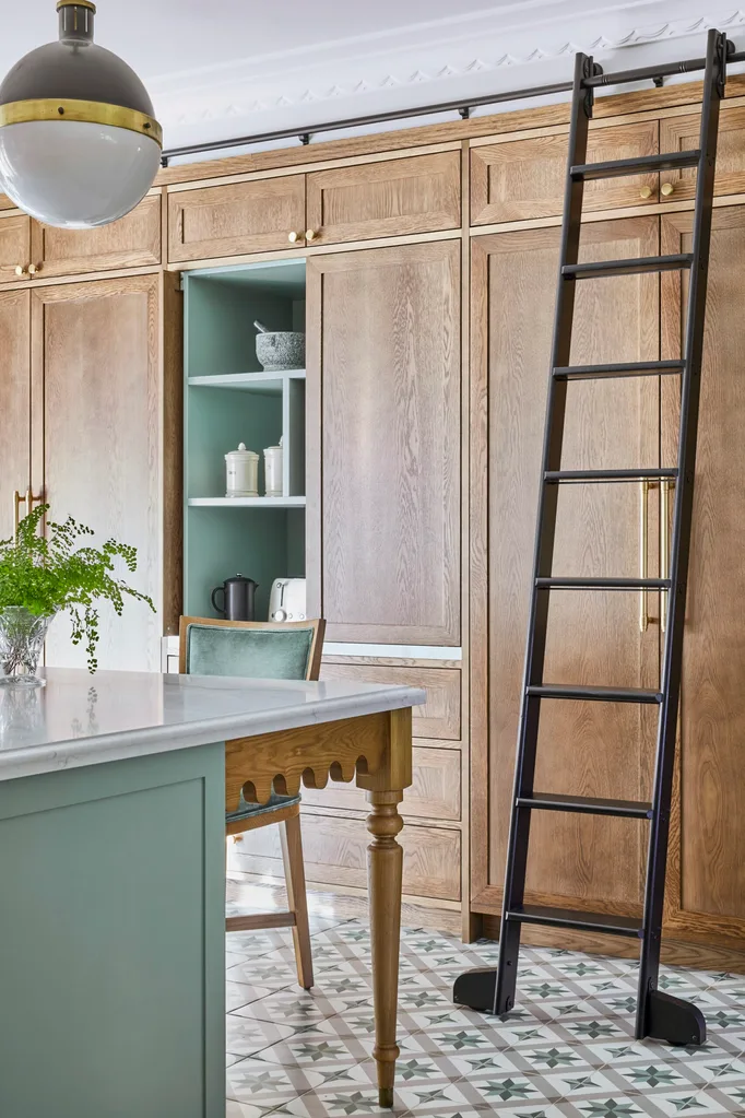 A green kitchen that was renovated with English style features to suit the 1921 build. A mint green finish is used on the kitchen island sides, with Calacatta Oro stone on the island benchtop. Wooden joinery reaches from floor to ceiling, with one cupboard open to show the green finish on the shelves. A black ladder can be wheeled across the joinery, to help the owners reach the highest shelves. The tiled flooring has a star pattern.