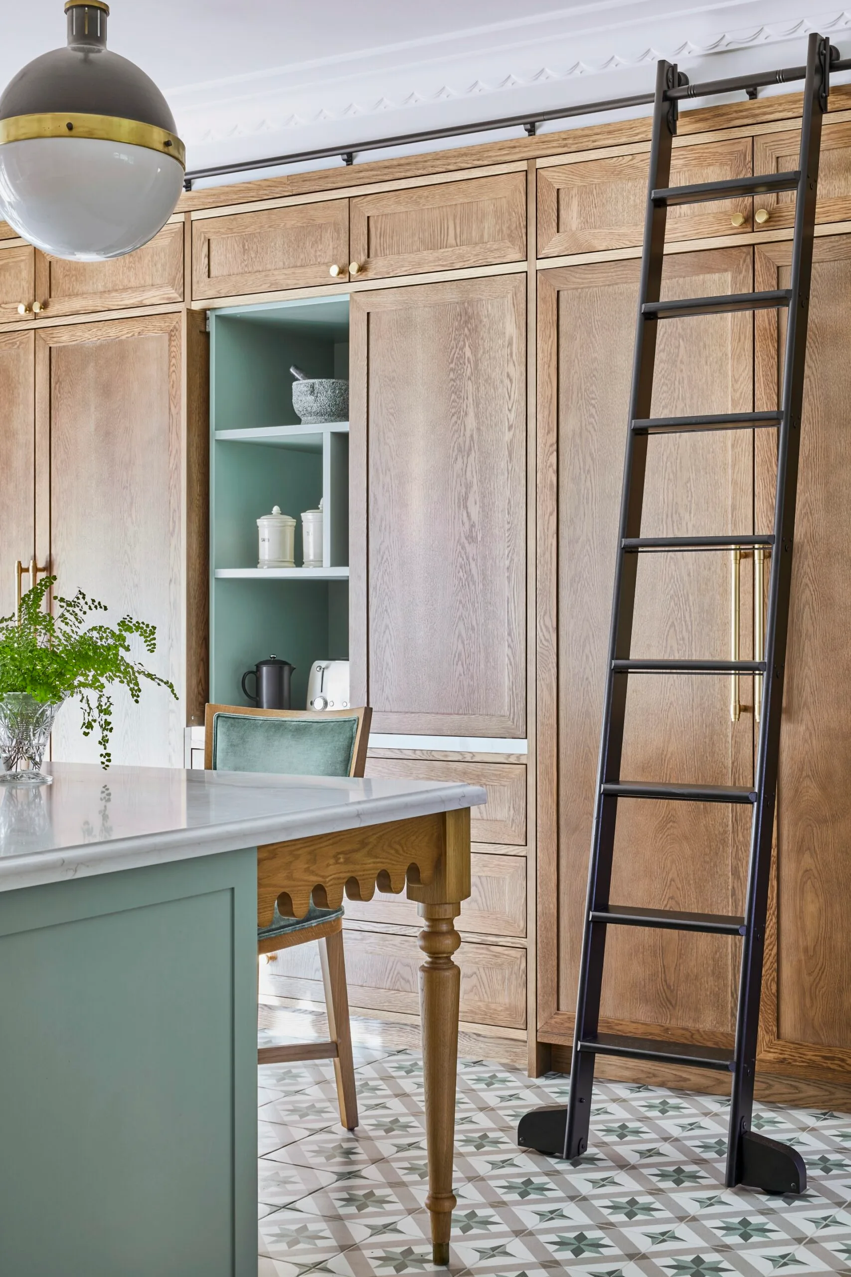 A green kitchen that was renovated with English style features to suit the 1921 build. A mint green finish is used on the kitchen island sides, with Calacatta Oro stone on the island benchtop. Wooden joinery reaches from floor to ceiling, with one cupboard open to show the green finish on the shelves. A black ladder can be wheeled across the joinery, to help the owners reach the highest shelves. The tiled flooring has a star pattern.