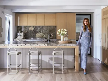 Bec Judd in the kitchen of her Arthurs Seat home, wearing a blue long sleeve top and matching long skirt with blue high heels. The kitchen has oak flooring, timber veneer joinery and marble benchtops.
