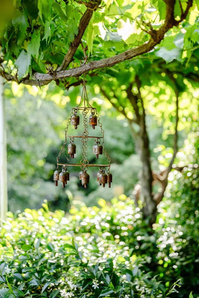 A patinaed wind charm that is shaped like a bell hangs from a tree, with bushes below.