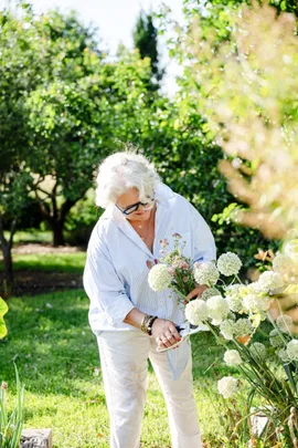 A woman wears white pants and a light blue buttoned shirt while using secateurs to cut flowers from her cottage garden.