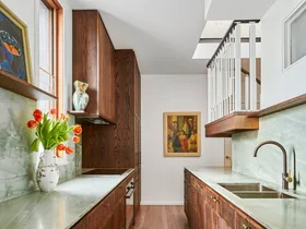 A galley kitchen with a light green quartzite that is used as the benchtop and the splashback, with underbench joinery made from hand-stained dark timber oak veneer. The tapware is also dark, in Antique Brass, with wooden flooring and framed paintings on the wall and an open shelf.