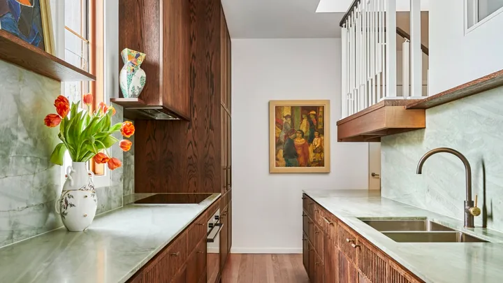 A galley kitchen with a light green quartzite that is used as the benchtop and the splashback, with underbench joinery made from hand-stained dark timber oak veneer. The tapware is also dark, in Antique Brass, with wooden flooring and framed paintings on the wall and an open shelf.