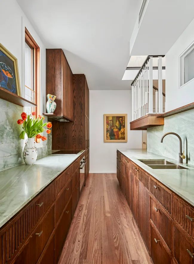 A galley kitchen with a light green quartzite that is used as the benchtop and the splashback, with underbench joinery made from hand-stained dark timber oak veneer. The tapware is also dark, in Antique Brass, with wooden flooring and framed paintings on the wall and an open shelf.