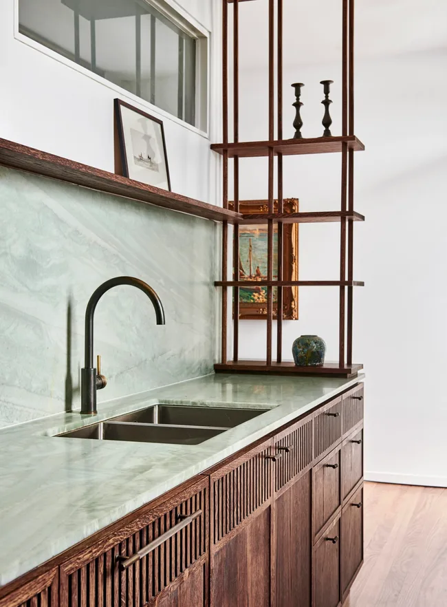 The end of a kitchen benchtop. Light green quartzite is used as the benchtop and the splashback, with underbench joinery made from hand-stained dark timber oak veneer. The tapware is also dark, in Antique Brass. A framed painting is hung on the wall, visible through open shelving that is made from the same dark veneer as the underbench joinery. A framed artwork is also on the open shelf above the splashback.