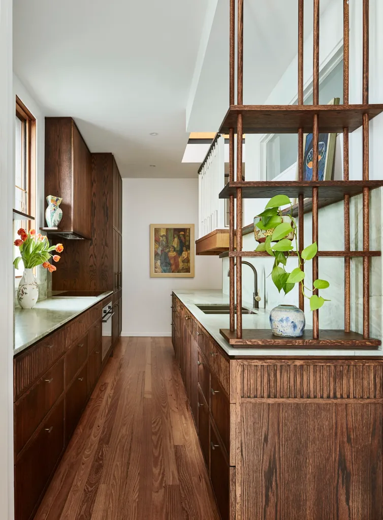 A galley kitchen with a light green quartzite that is used as the benchtop and the splashback, with underbench joinery made from hand-stained dark timber oak veneer. The tapware is also dark, in Antique Brass. Timber floorboards complement the joinery. In the foreground, open shelves start at the benchtop and reach upwards, with two ceramic pots positioned on them. One has a devil's ivy plant gently reaching down. Paintings are on the open shelf and the wall in the background.