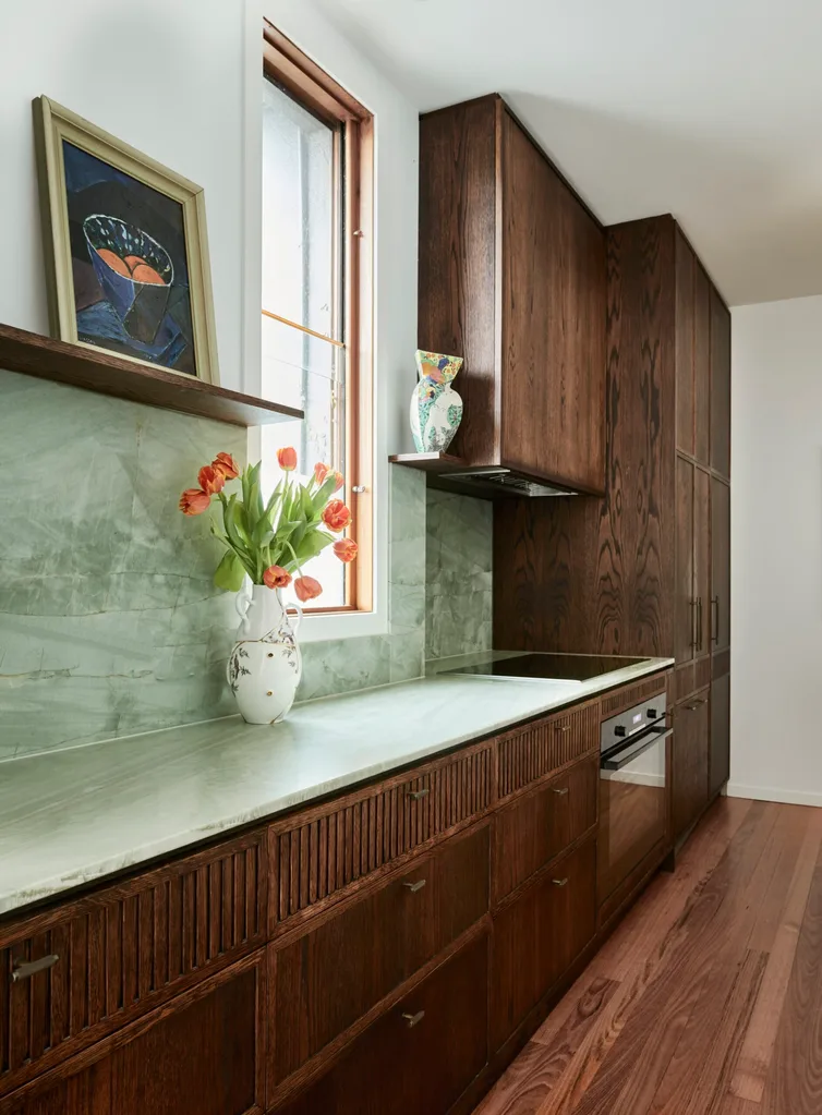 A galley kitchen with a light green quartzite that is used as the benchtop and the splashback, with underbench joinery made from hand-stained dark timber oak veneer. This joinery continues past the benchtop, creating floor-to-ceiling cupboards with Antique Brass handles. A vase on the benchtop holds orange tulips, while two open shelves showcase a framed painting and a ceramic piece of art.
