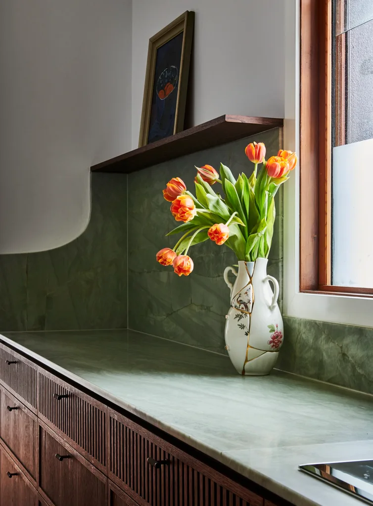 This kitchen has a light green quartzite that is used as the benchtop and the splashback, with underbench joinery made from hand-stained dark timber oak veneer. A vase on the benchtop holds orange tulips. An open shelf in the background has a framed painting positioned on it.