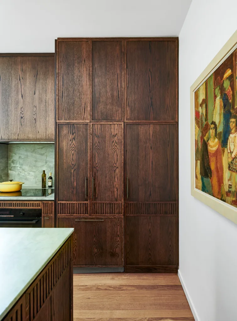 A galley kitchen with a light green quartzite that is used as the benchtop and the splashback, with underbench joinery made from hand-stained dark timber oak veneer. This joinery continues past the benchtop, creating floor-to-ceiling cupboards with Antique Brass handles. The wall is finished in Dulux Natural White with a framed painting placed there.