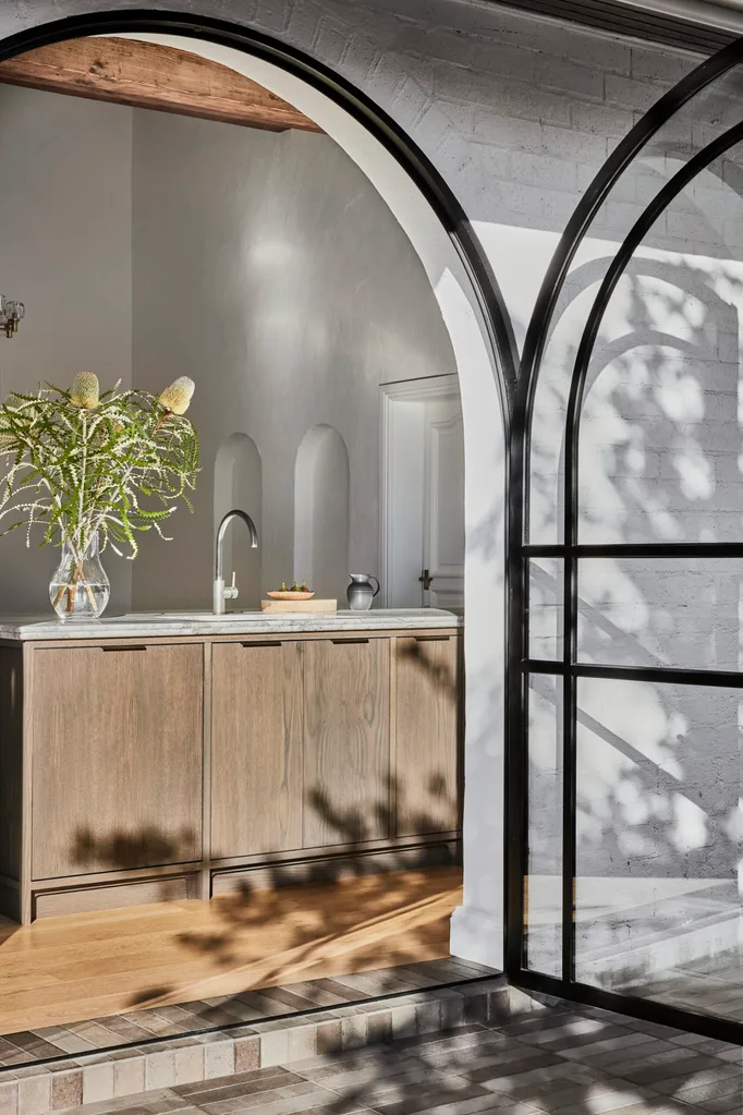In the foreground a black steel and glass French door is open, revealing an open plan kitchen inside. Oak veneer joinery was used on the kitchen island, with curved tapware above the kitchen basin. To the left, native plants are positioned within a glass vase.