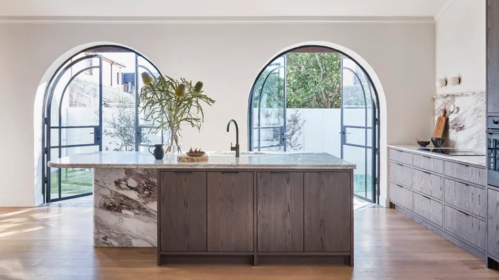 A kitchen with oak timber floorboards, oak veneer joinery, structural timber beams along the white ceiling, marble benchtops, marble splashbacks and two arched French doors in glass and black steel. The kitchen is open plan with a sizable island in the middle.