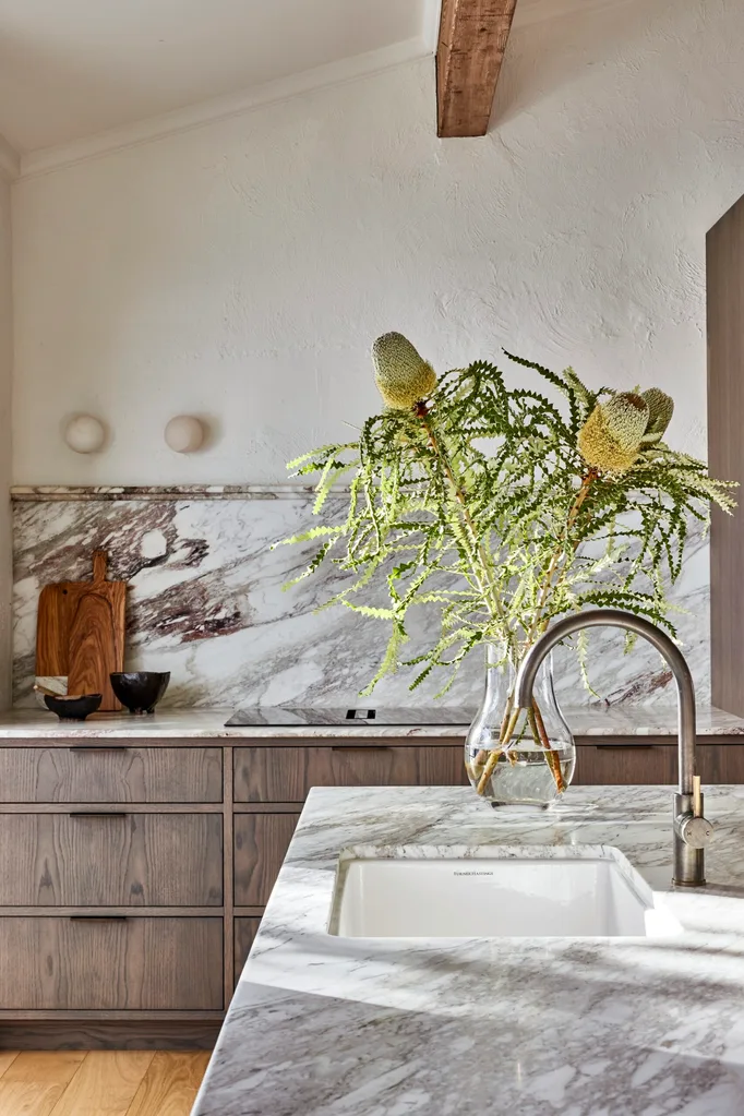 A kitchen with underbench oak veneer joinery, marble benchtops, marble splashbacks and an arched French doors in glass and black steel. A native plant sits in a glass on the kitchen island, to the right of the photo. Tapware is positioned above a basin, and has an arched design.