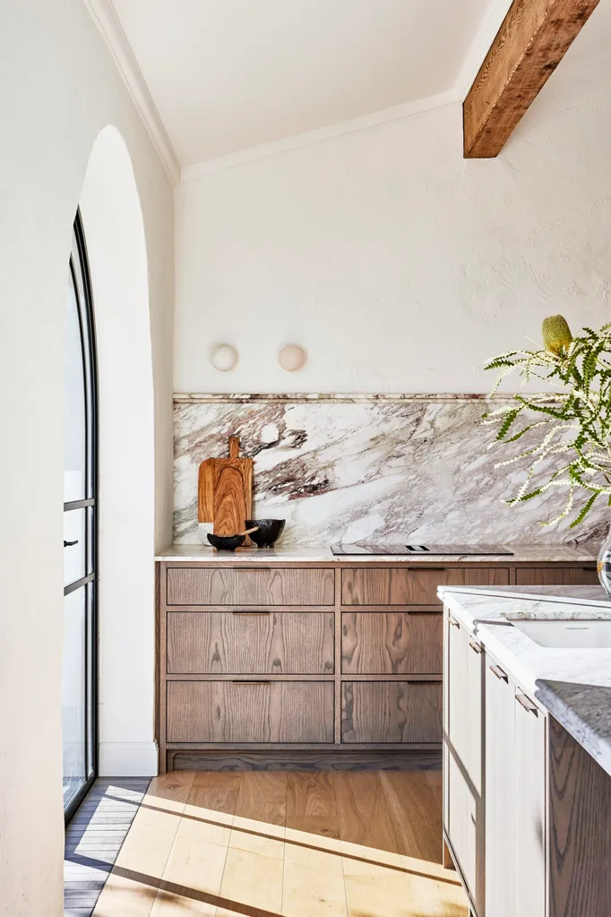 A kitchen with underbench oak veneer joinery, marble benchtops, marble splashbacks and an arched French doors in glass and black steel. A native plant sits in a glass on the kitchen island, to the right of the photo.