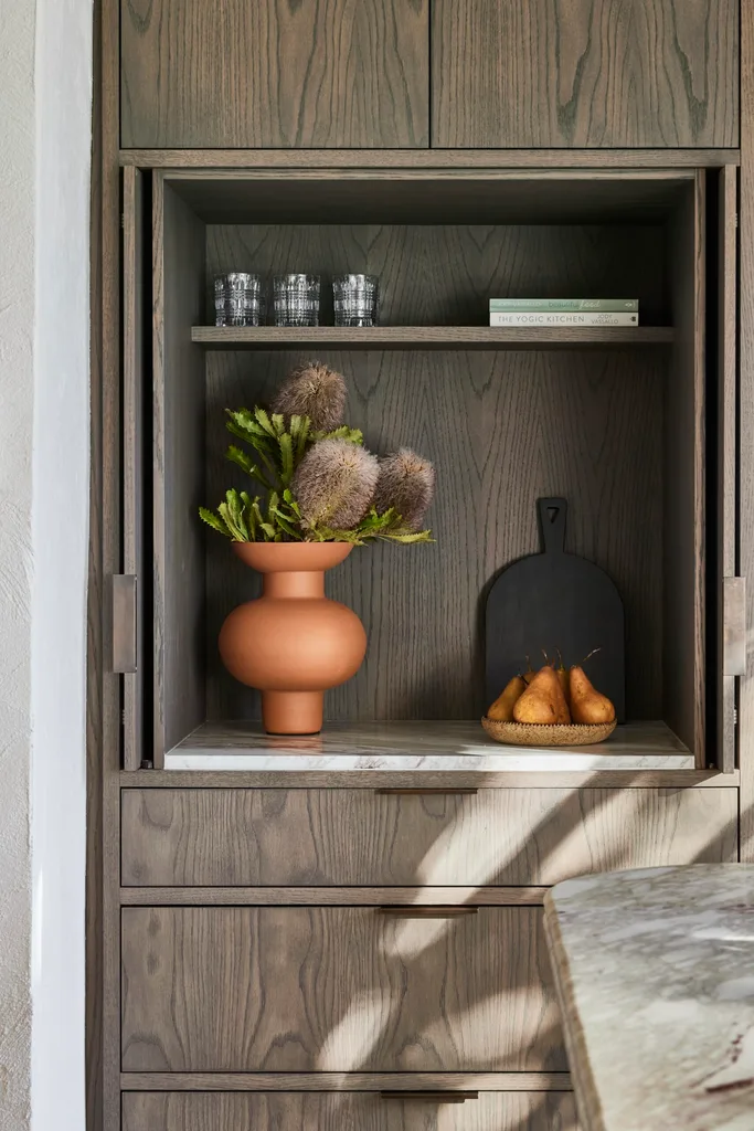 A close up shot of oak veneer joinery. Drawers are positioned underneath a cupboard, which is open to reveal an orange vase with native plants inside, a platter of fruit and a black chopping board.