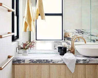 A modern bathroom with timber joinery underneath a marble benchtop, a round white basin and gold hardware.