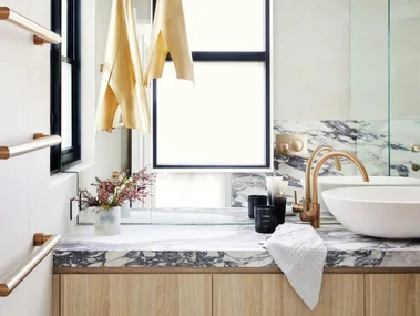 A modern bathroom with timber joinery underneath a marble benchtop, a round white basin and gold hardware.