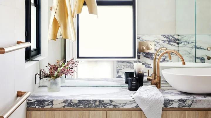 A modern bathroom with timber joinery underneath a marble benchtop, a round white basin and gold hardware.