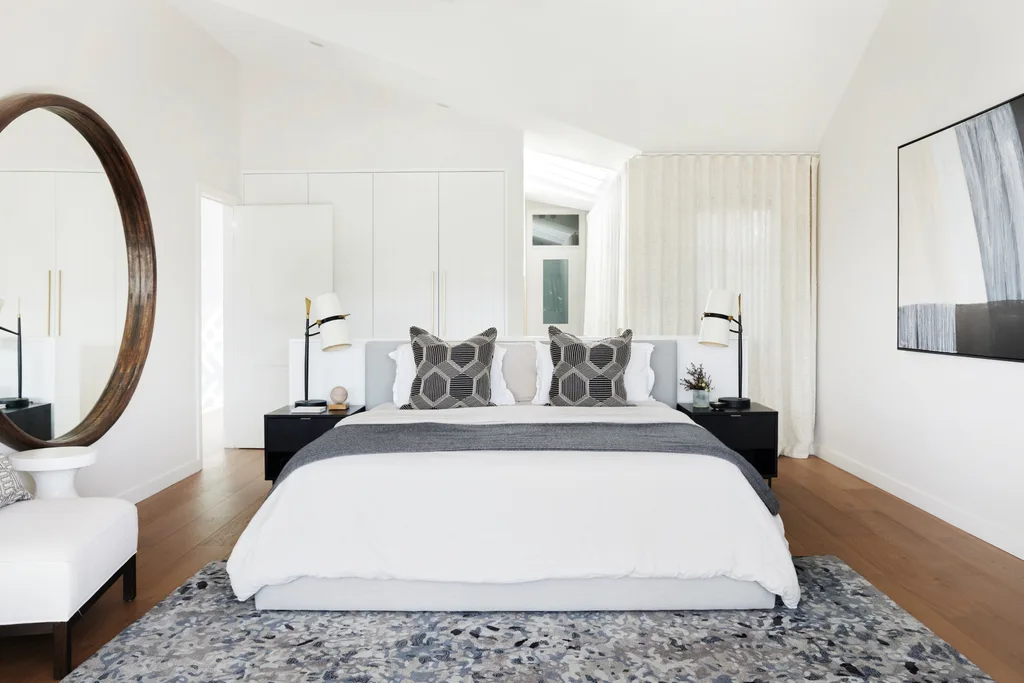 A modern bedroom with monochromatic bed linen, a circular mirror on the wall and two black bedside tables with black and white table lamps on top