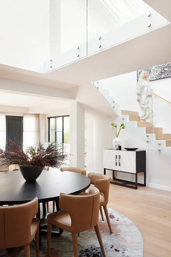 A modern white interior with a round dining table, stairs leading up to the second floor and a woman walking down the stairs