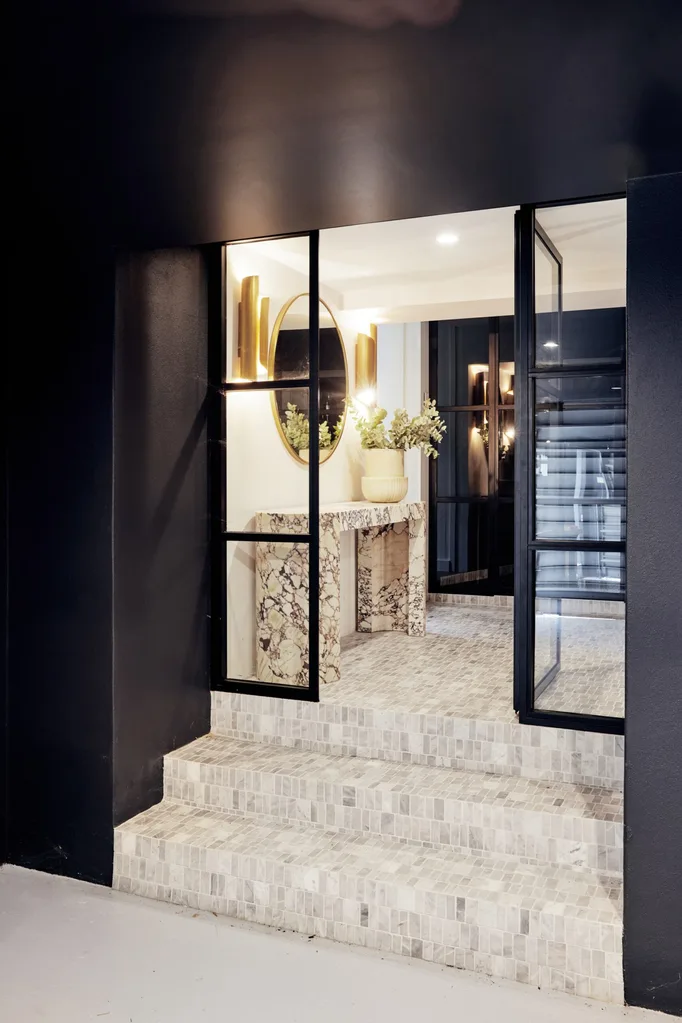 A black facade with three tiled steps leading to an entranceway, which has a mirror, two gold light fittings and a marble console table.