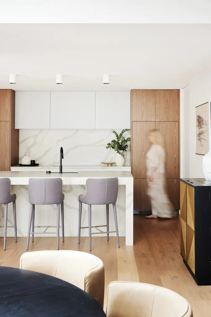 A kitchen with white benchtops, a white island, white cabinetry above the benchtop then timber cabinetry on either side. A woman walks in front of the timber cabinetry.