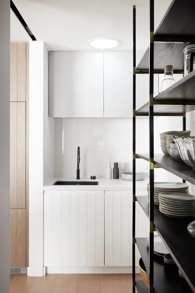A monochromatic butler's pantry with white splashback, benchtop and cabinetry near black open shelving and modern black tapware.