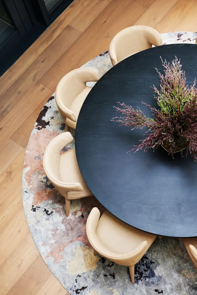 A round black dining table with greenery in the middle, surrounded by cream dining chairs atop a circular rug