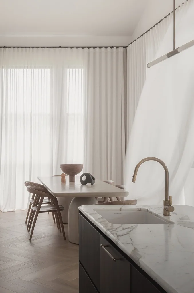 Neutral dining room featuring timber chairs, a marble kitchen benchtop, brass tapware and sheer curtains