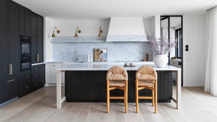 A kitchen with a sizable kitchen island as the centrepiece. Carrara marble is the splashback and benchtop, with island sides that are finished in Porter's Paints Black Cockatoo. Bar stools are timber with woven detailing on the seat and cushioned back. The white rangehood cover has a strip of carrara marble around the base. Wall lights are positioned next to the rangehood, with an antique brass finish. A hidden bar is concealed in the joinery to the left.