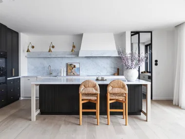 A kitchen with a sizable kitchen island as the centrepiece. Carrara marble is the splashback and benchtop, with island sides that are finished in Porter's Paints Black Cockatoo. Bar stools are timber with woven detailing on the seat and cushioned back. The white rangehood cover has a strip of carrara marble around the base. Wall lights are positioned next to the rangehood, with an antique brass finish. A hidden bar is concealed in the joinery to the left.
