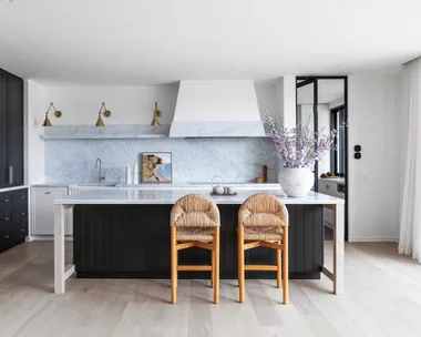 A kitchen with a sizable kitchen island as the centrepiece. Carrara marble is the splashback and benchtop, with island sides that are finished in Porter's Paints Black Cockatoo. Bar stools are timber with woven detailing on the seat and cushioned back. The white rangehood cover has a strip of carrara marble around the base. Wall lights are positioned next to the rangehood, with an antique brass finish. A hidden bar is concealed in the joinery to the left.
