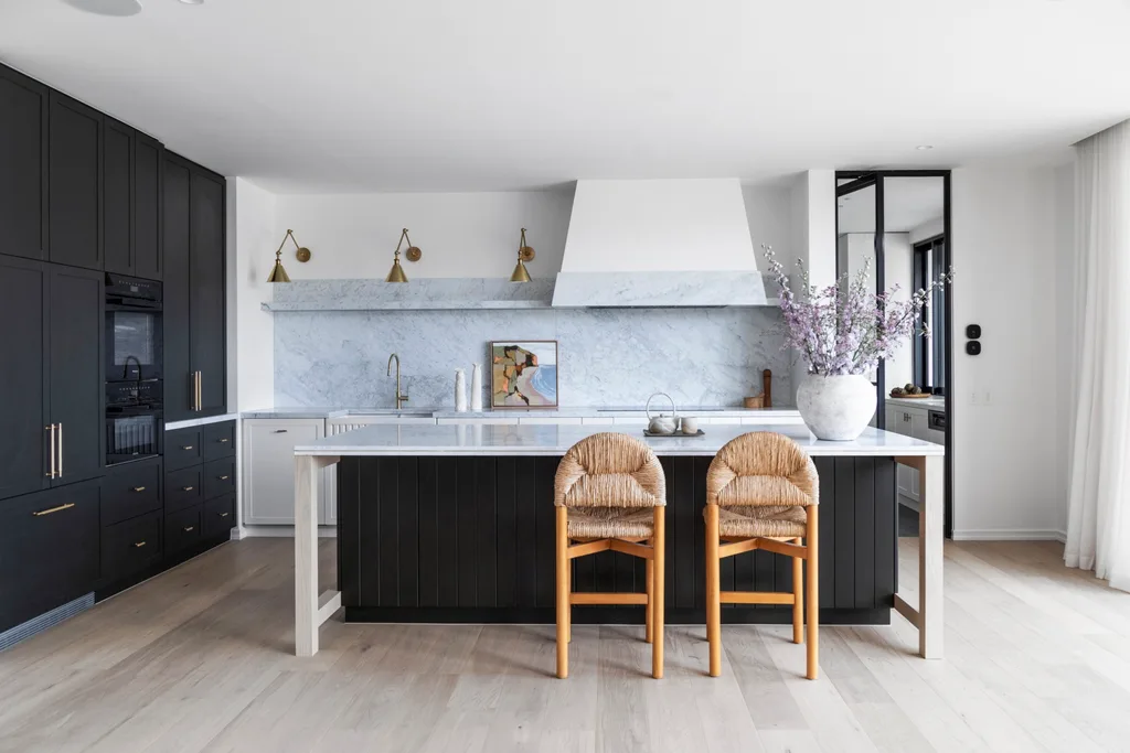 A kitchen with a sizable kitchen island as the centrepiece. Carrara marble is the splashback and benchtop, with island sides that are finished in Porter's Paints Black Cockatoo. Bar stools are timber with woven detailing on the seat and cushioned back. The white rangehood cover has a strip of carrara marble around the base. Wall lights are positioned next to the rangehood, with an antique brass finish. A hidden bar is concealed in the joinery to the left.