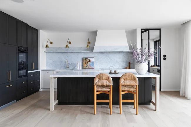 A kitchen with a sizable kitchen island as the centrepiece. Carrara marble is the splashback and benchtop, with island sides that are finished in Porter's Paints Black Cockatoo. Bar stools are timber with woven detailing on the seat and cushioned back. The white rangehood cover has a strip of carrara marble around the base. Wall lights are positioned next to the rangehood, with an antique brass finish. A hidden bar is concealed in the joinery to the left.