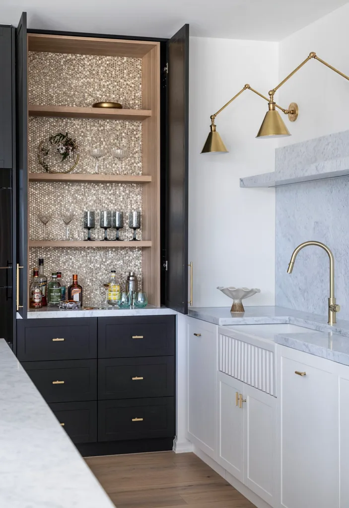 Joinery doors open to reveal a hidden bar, with golden hexagonal mosaic tiles on the back wall, timber open shelves and plenty of glassware, spirits and cocktail shakers.