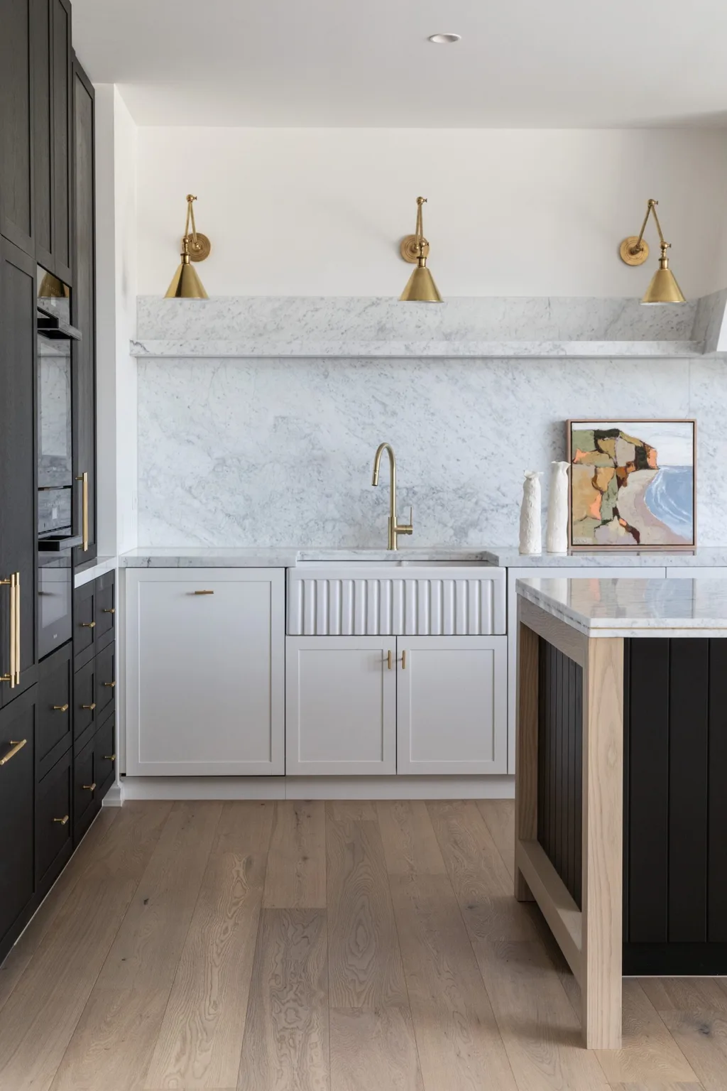 Carrara marble is the splashback and benchtop in this kitchen, with island sides that are finished in Porter's Paints Black Cockatoo. Wall lights are positioned above the splashback, with an antique brass finish. A hidden bar is concealed in the joinery to the left. A painting is on the benchtop beside the sink.