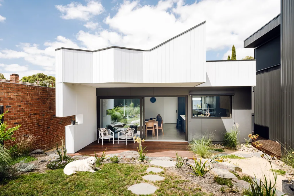The rear exterior of a certified passive house. A dog sits on the grass in the backyard. Sliding doors are open, revealing an open plan kitchen, dining and living area.