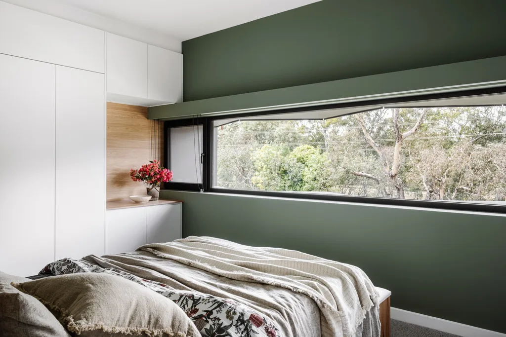 A bedroom with one feature wall painted green. A long, ribbon window stretches across this wall, showing views of surrounding bushland.