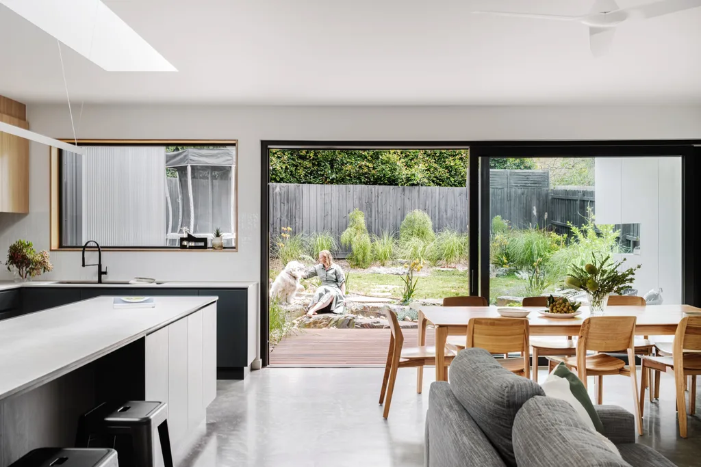 An open plan kitchen, dining and living space that leads to a backyard. A woman in a button up dress sits outside with a dog.