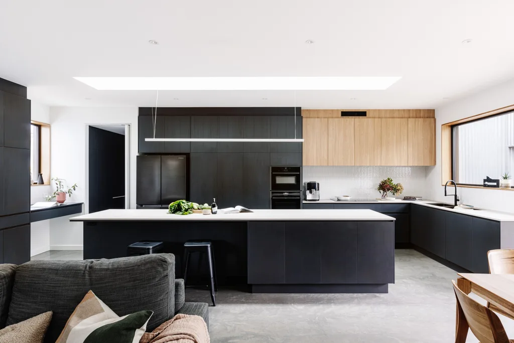 A kitchen in a certified passive house, with a sizable kitchen island, a study nook to one side and a large window looking out to the backyard. Glossy white tiles form the splashback with joinery made of Laminex products.