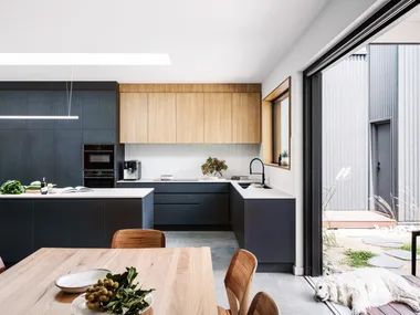 A kitchen in a certified passive house, with a sizable kitchen island and a large window looking out to the backyard. Glossy white tiles form the splashback with joinery made of Laminex products. A dog lays down at the entrance to the yard.